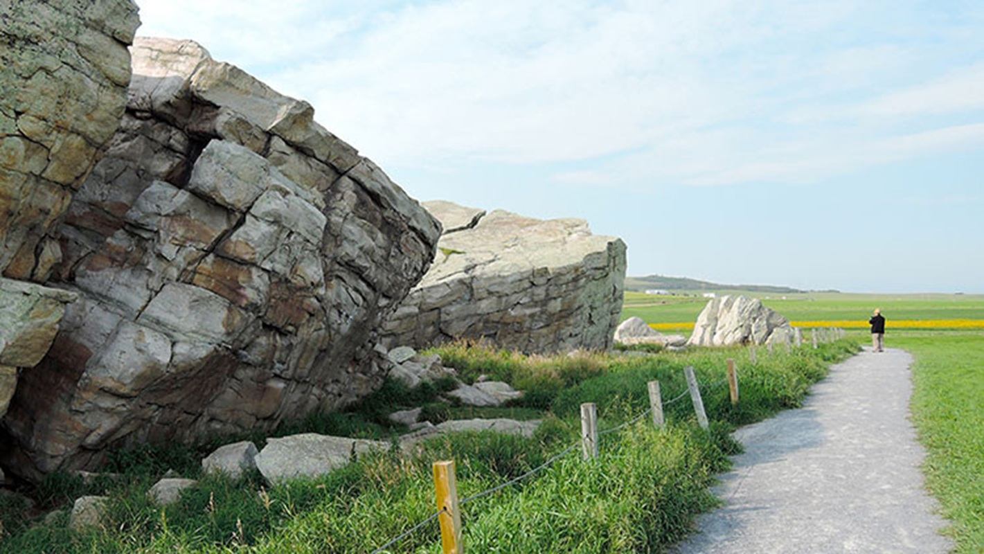 Large rock formations beside a gravel path through open green fields under blue sky.