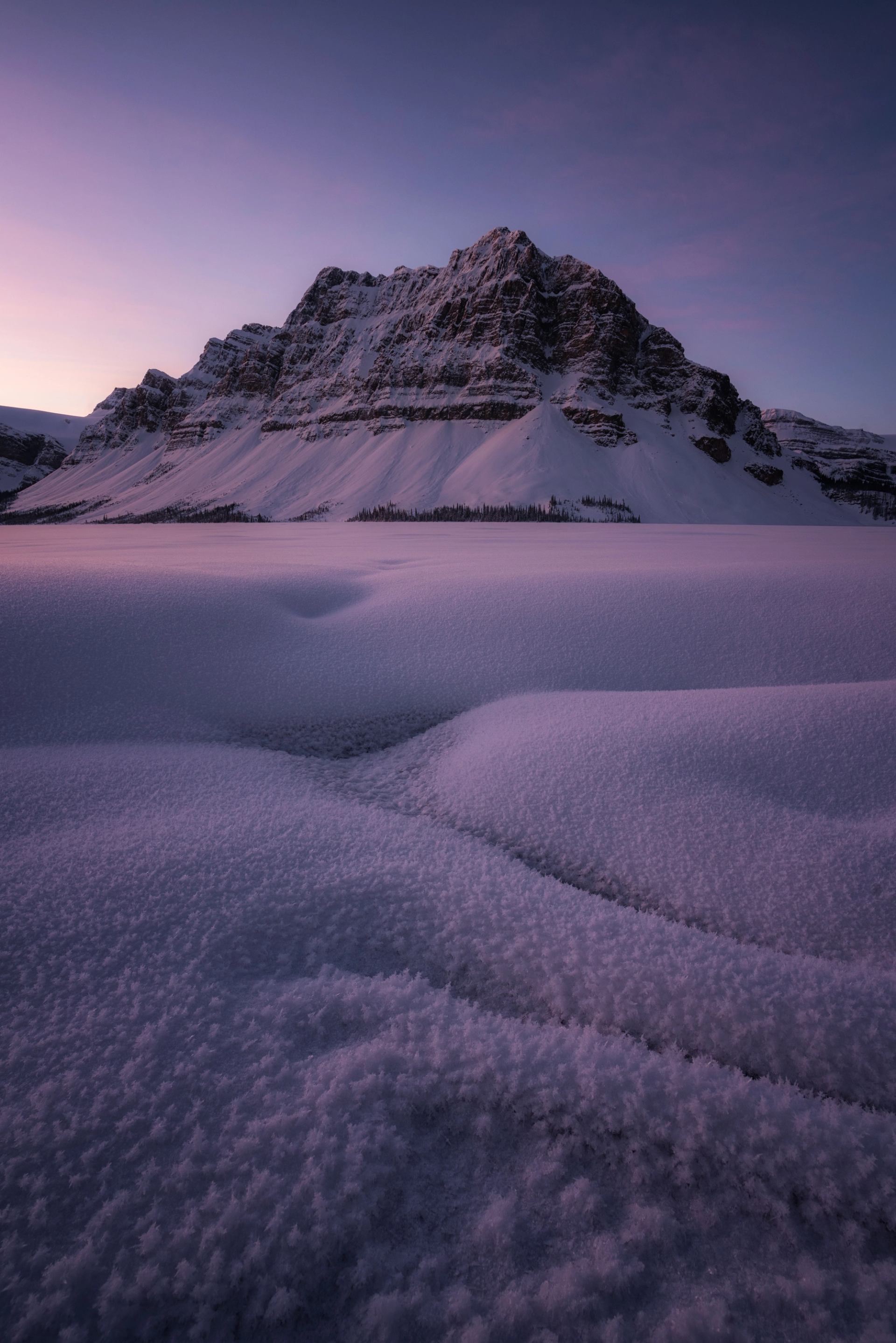 Snow-covered mountain at sunset with soft pink and purple hues across the frozen landscape.