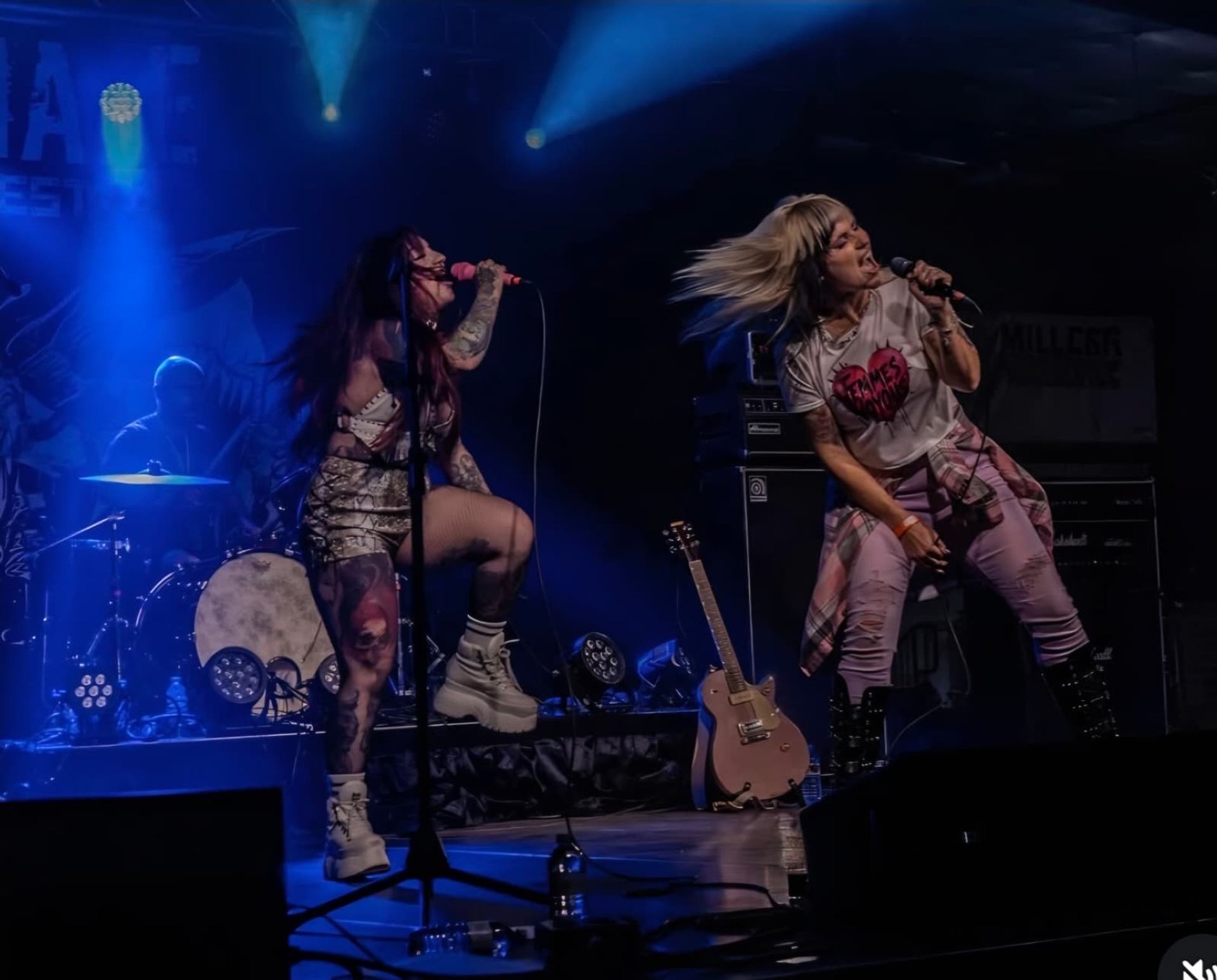 Two singers perform with microphones on a blue-lit stage beside a drummer and guitar.