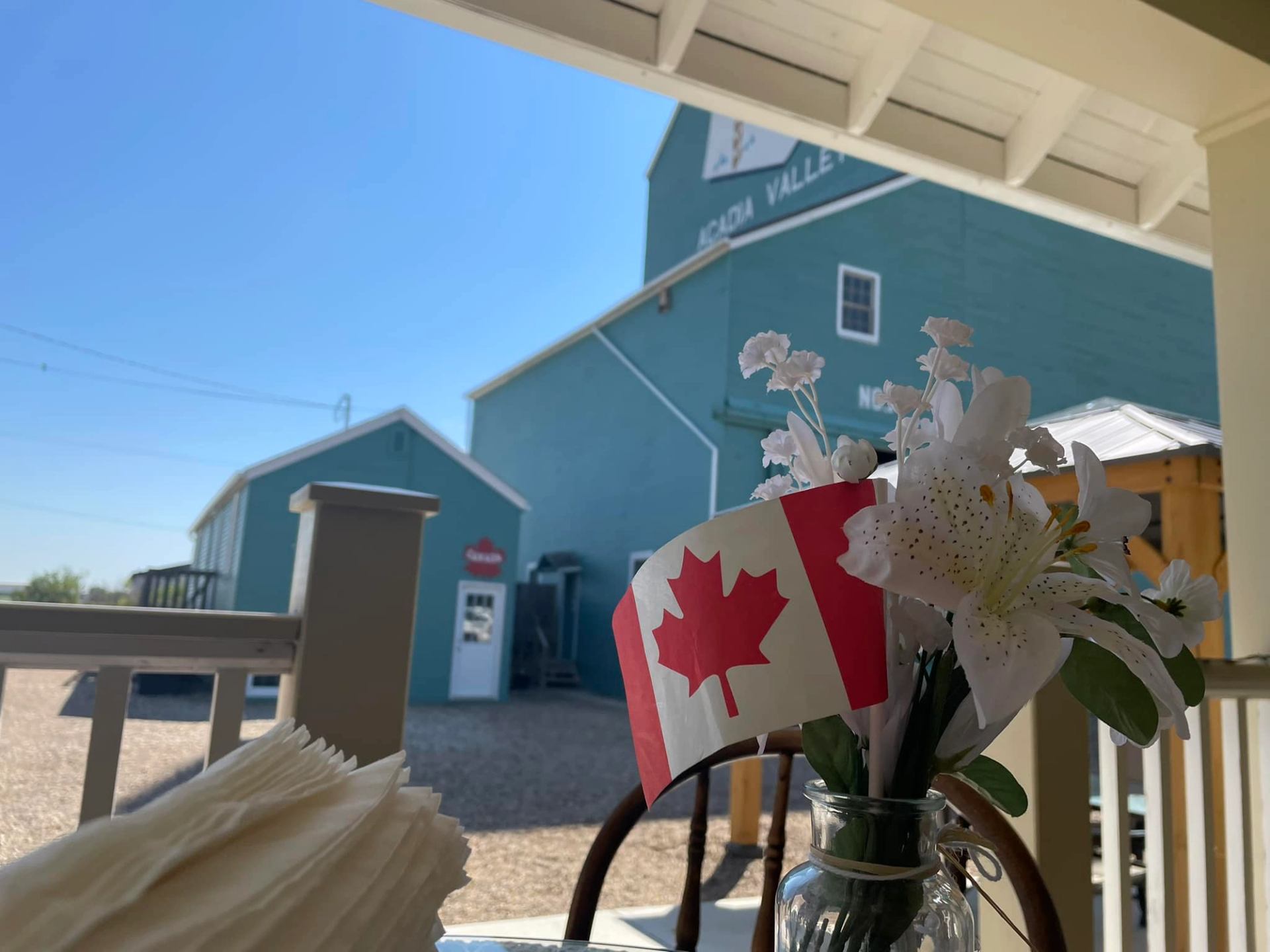 Canadian flag and white flowers in vase on porch, blue farm buildings behind.