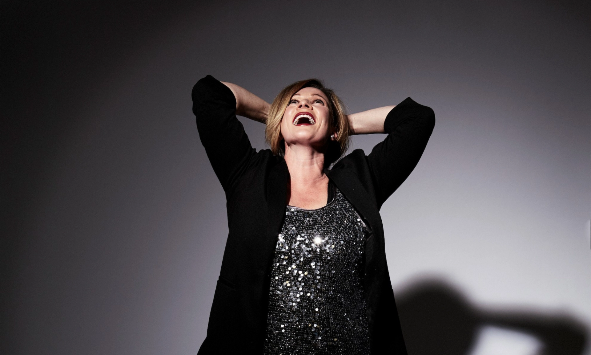 Person in black blazer and sequined top stands with hands behind head, smiling.