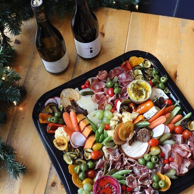 Charcuterie tray with meats, cheeses, vegetables and fruit beside wine bottles on wooden table.