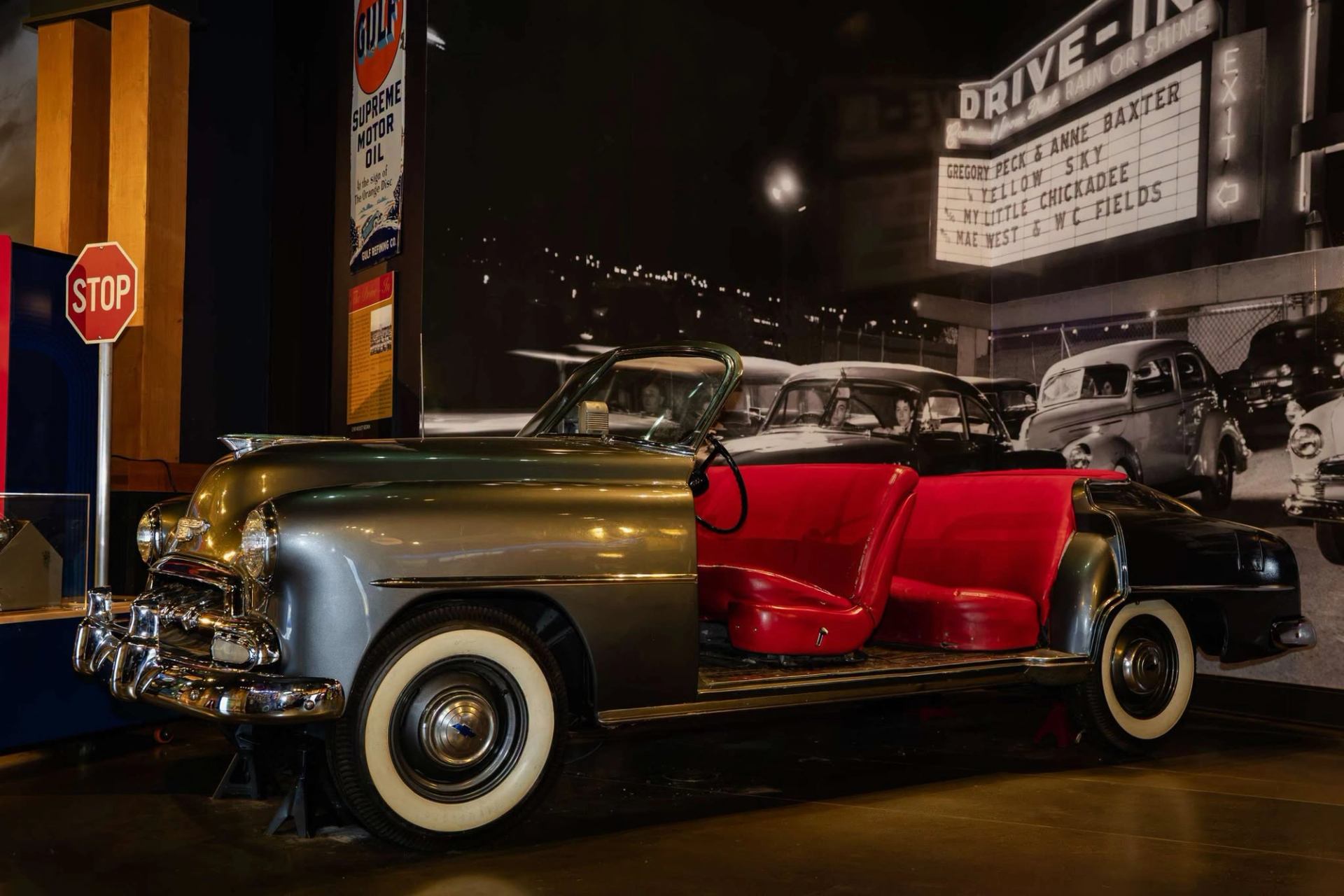 Vintage convertible car with red interior displayed indoors beside drive-in theatre backdrop.