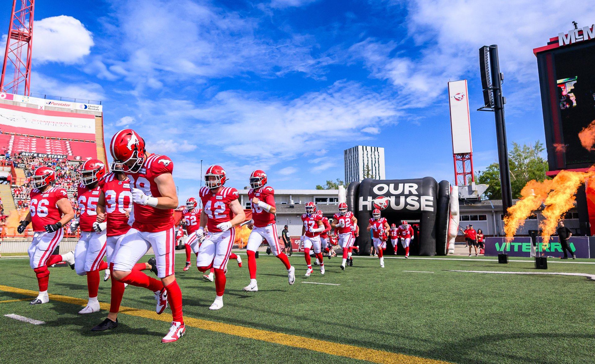 Football players in red uniforms run onto the field beside bursts of flame.