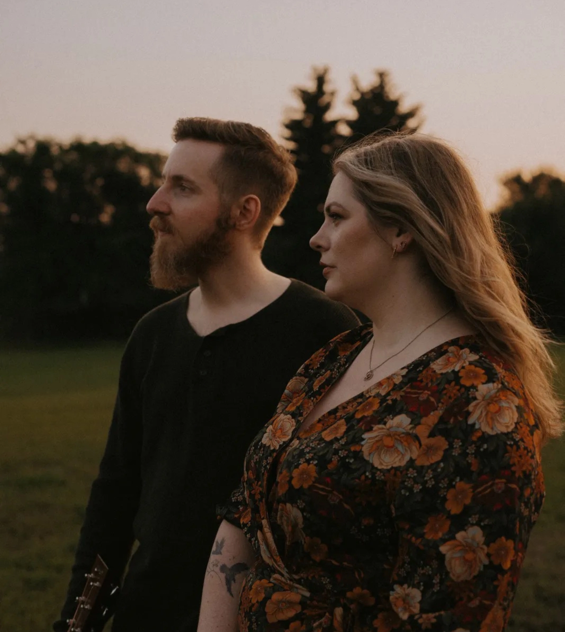 Two musicians stand in an open field at sunset, looking into the distance with trees visible in the background.