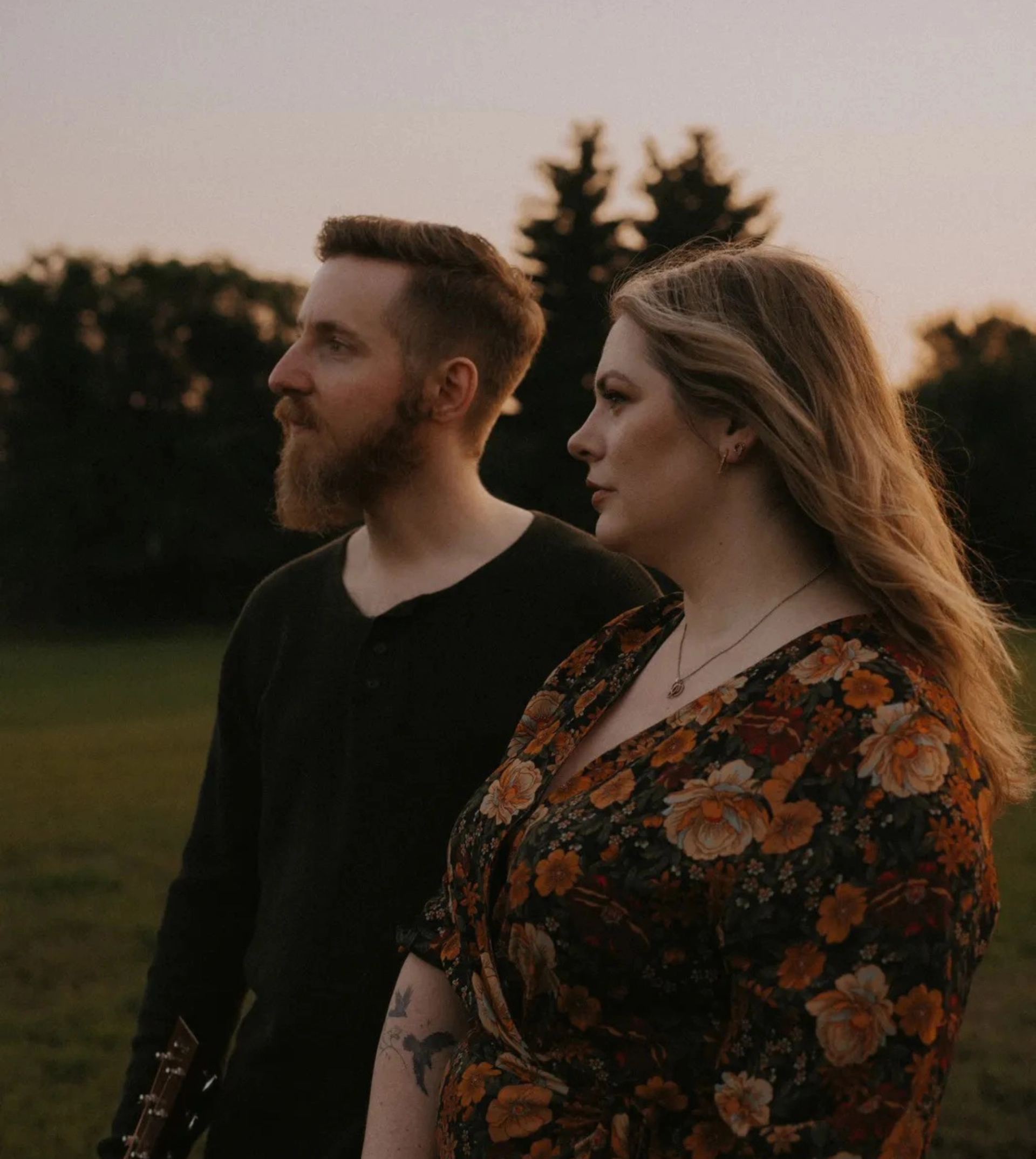Two musicians stand in an open field at sunset, looking into the distance with trees visible in the background.