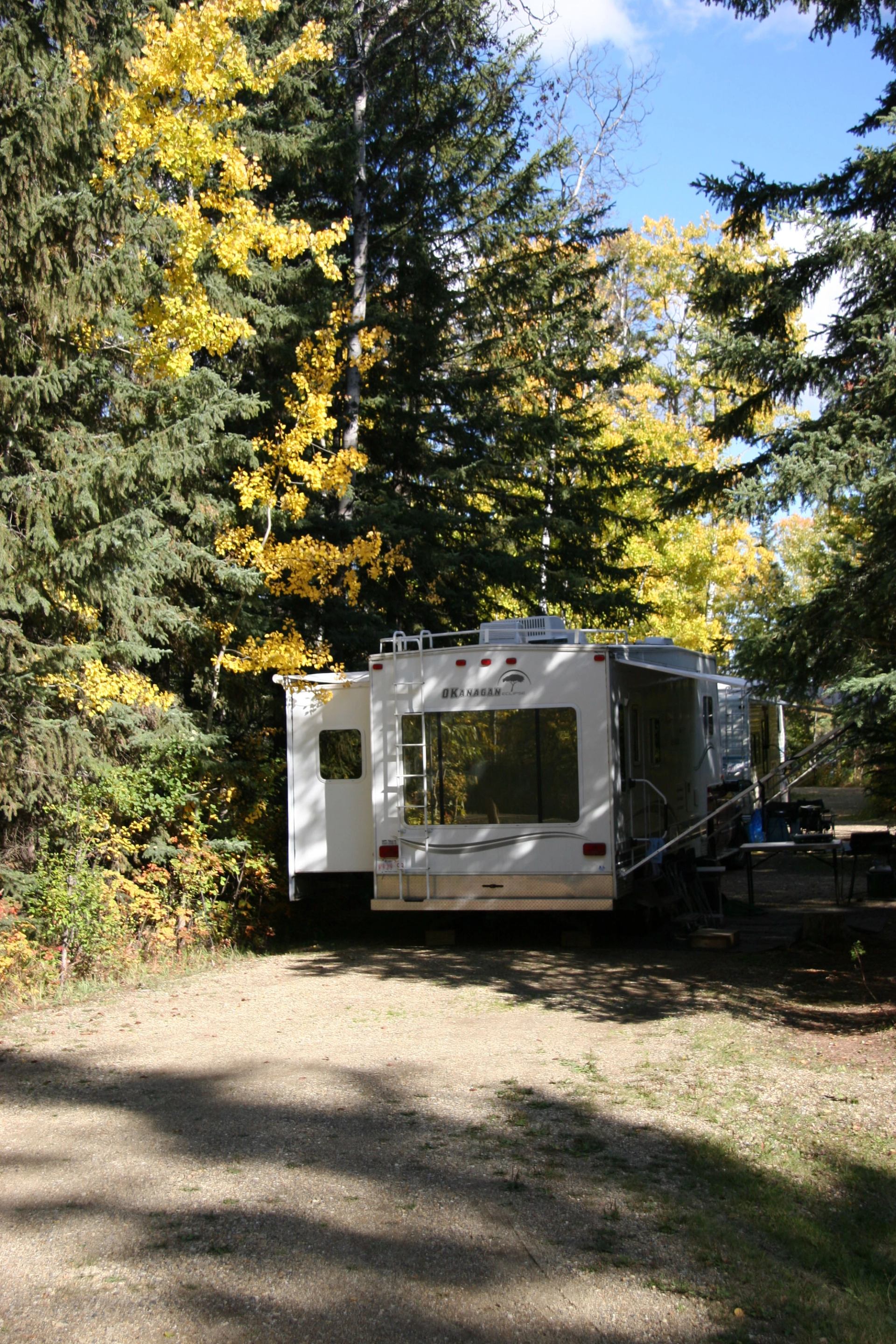 White RV parked on forest path surrounded by autumn trees and sunlight.