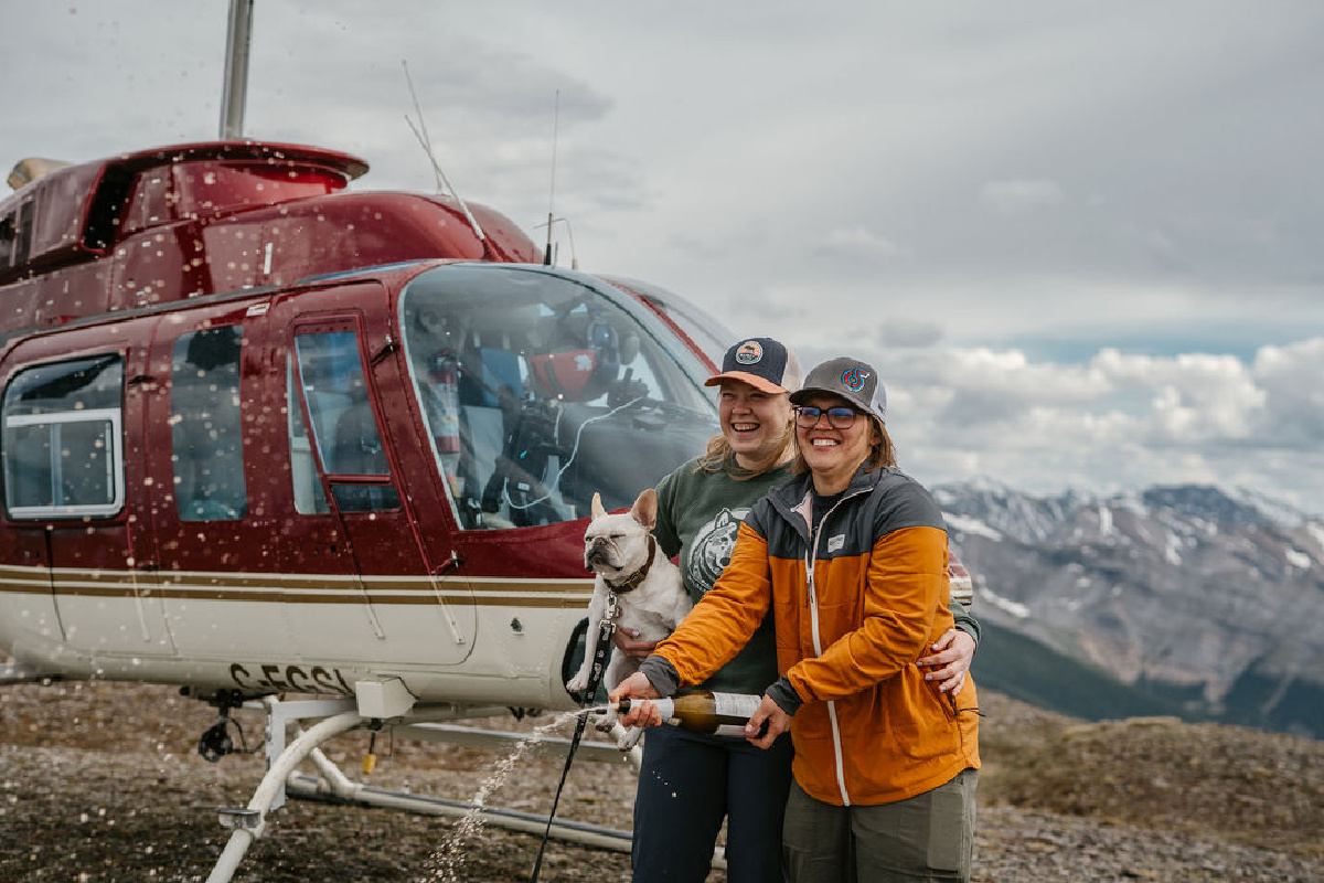 Two people smile with their dog beside a helicopter on a mountain ridge.