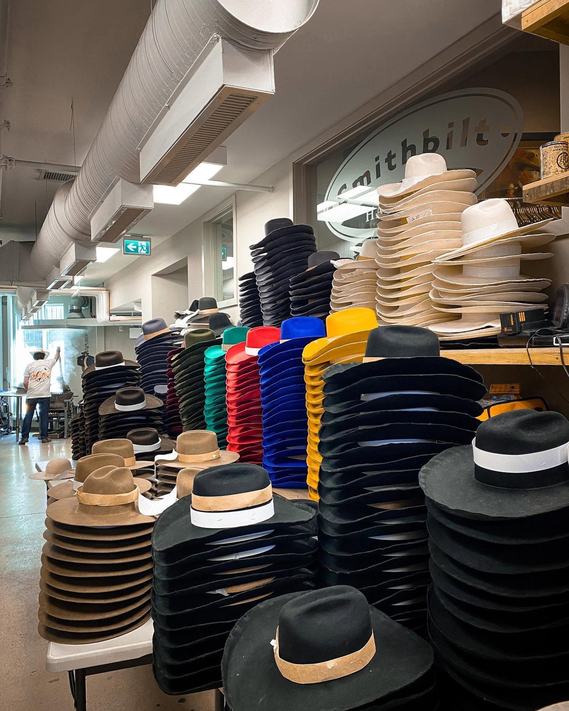 Stacks of colourful cowboy hats fill a workshop, with ventilation ducts overhead.