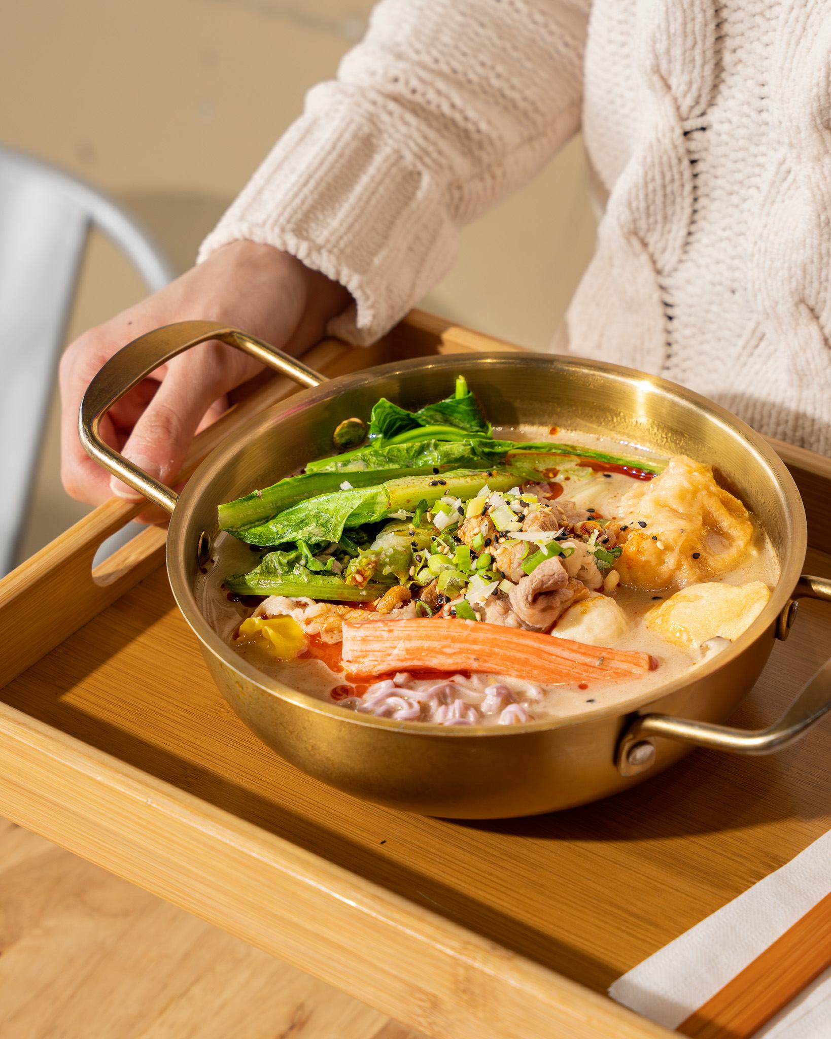 Golden pot with greens, tofu, crab sticks, and veggies on a wooden tray, held by a person.