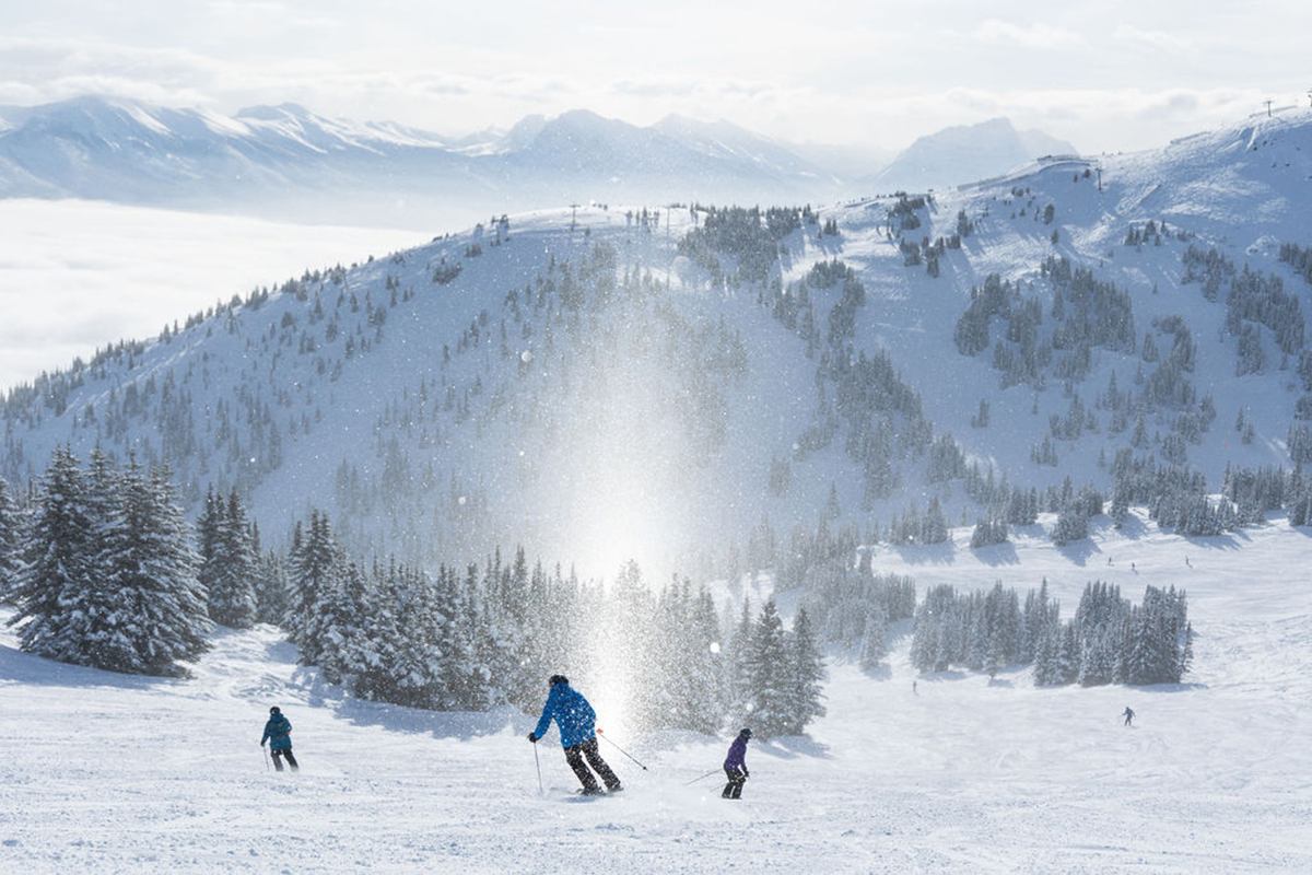 Three skiers going down a blue rated slope with a Mountain range in the background.
