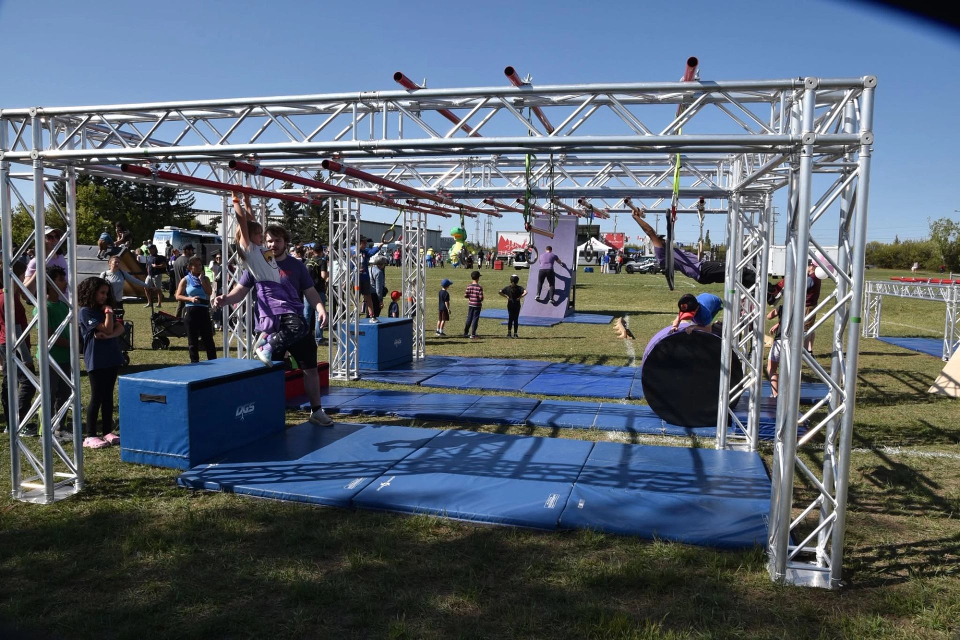 Outdoor obstacle course with children climbing metal bars and adults supervising on blue mats.