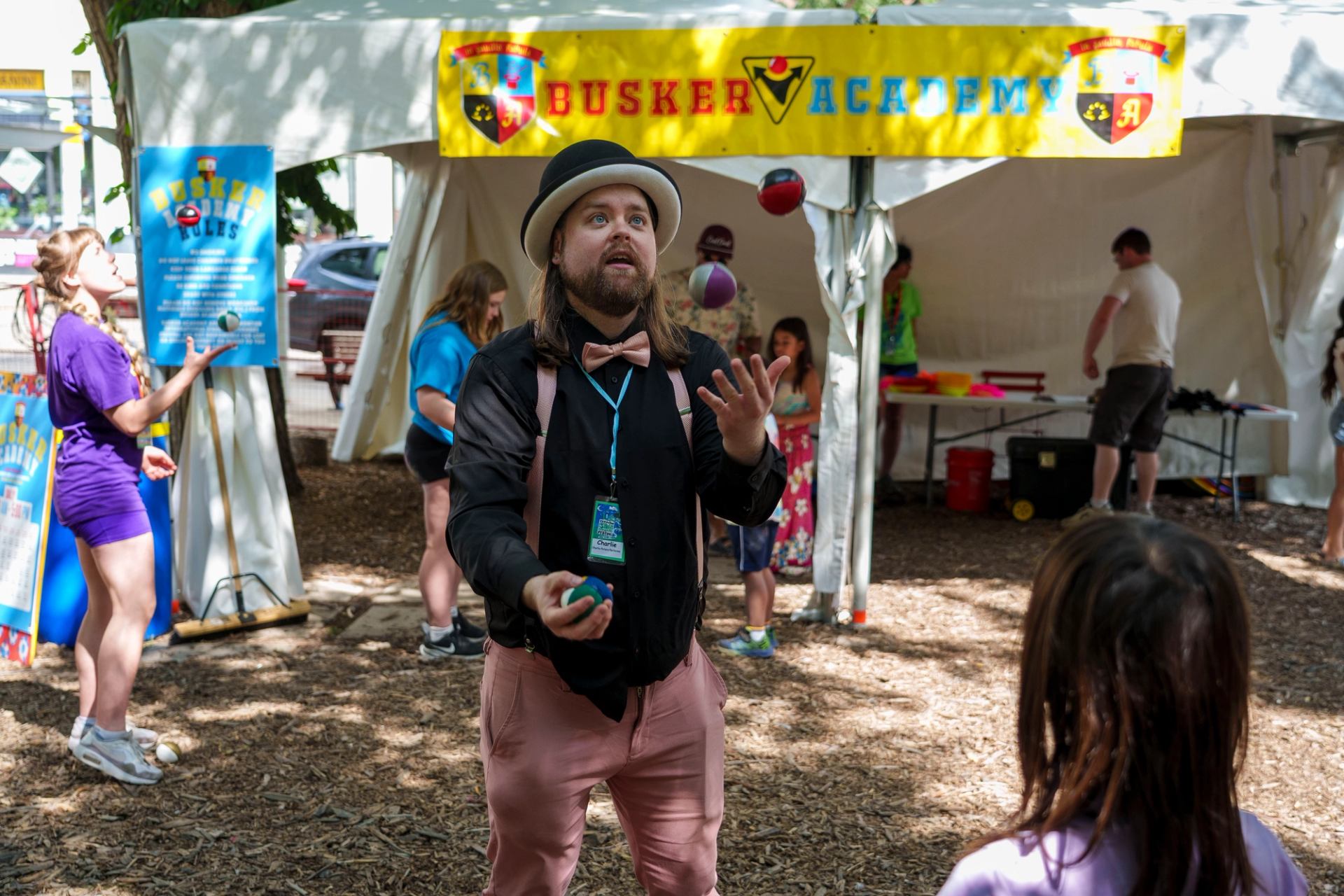 Street performer in hat juggles colourful balls at Busker Academy festival tent.