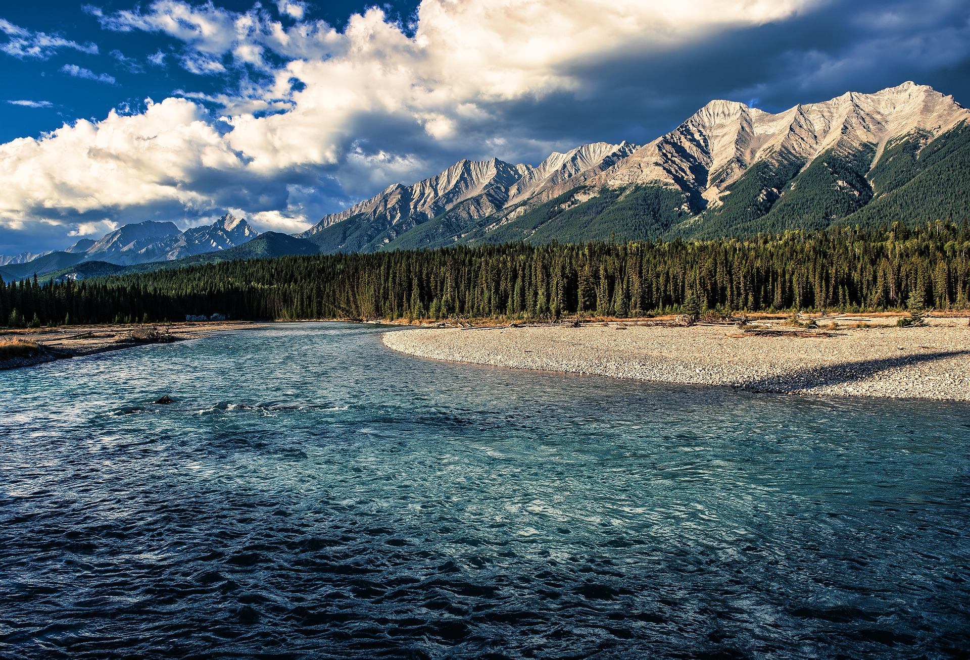Wide blue river curves past rocky shore, dense evergreen forest and rugged mountains.