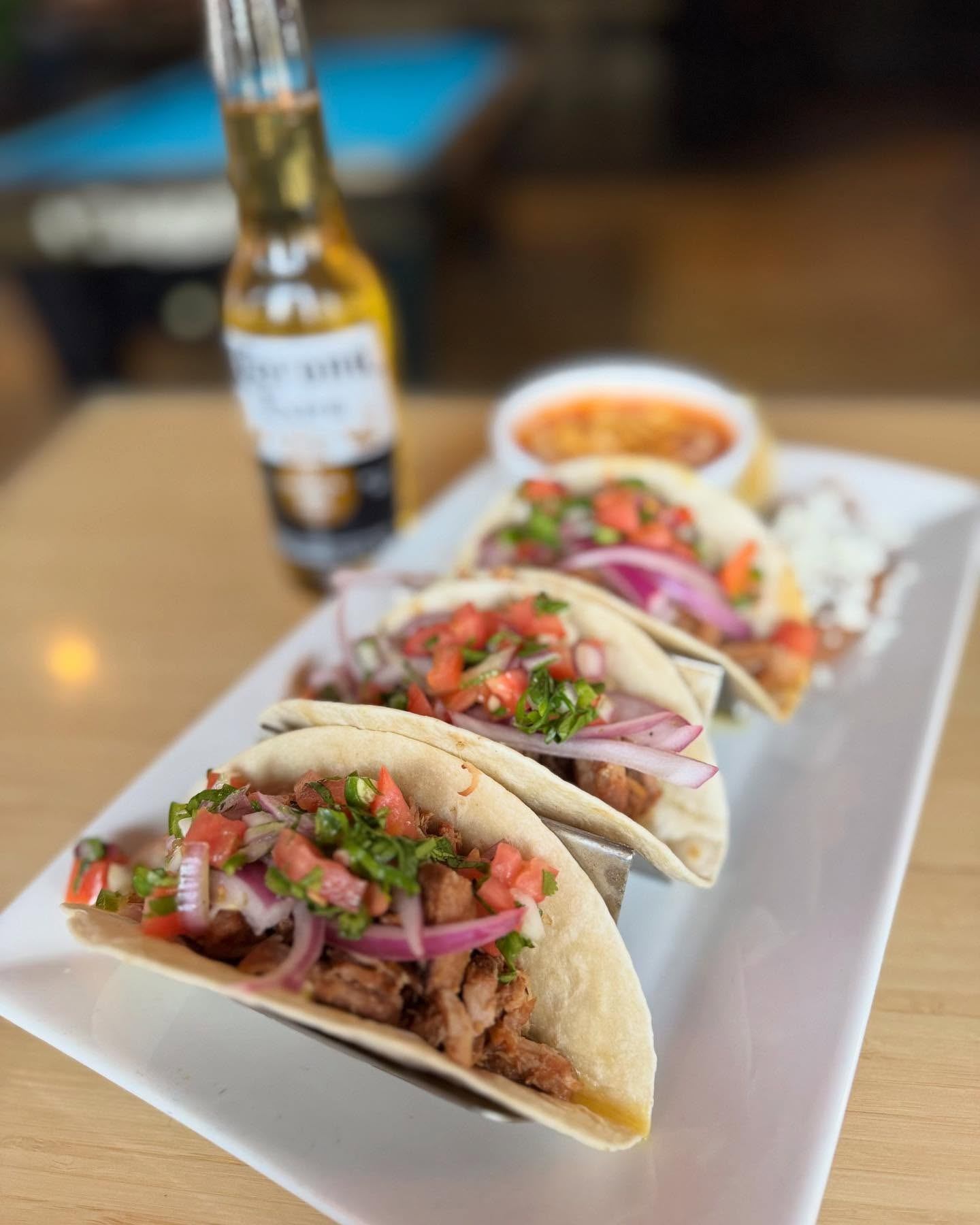 Tacos topped with tomatoes, red onion and cilantro beside salsa and a beer bottle.
