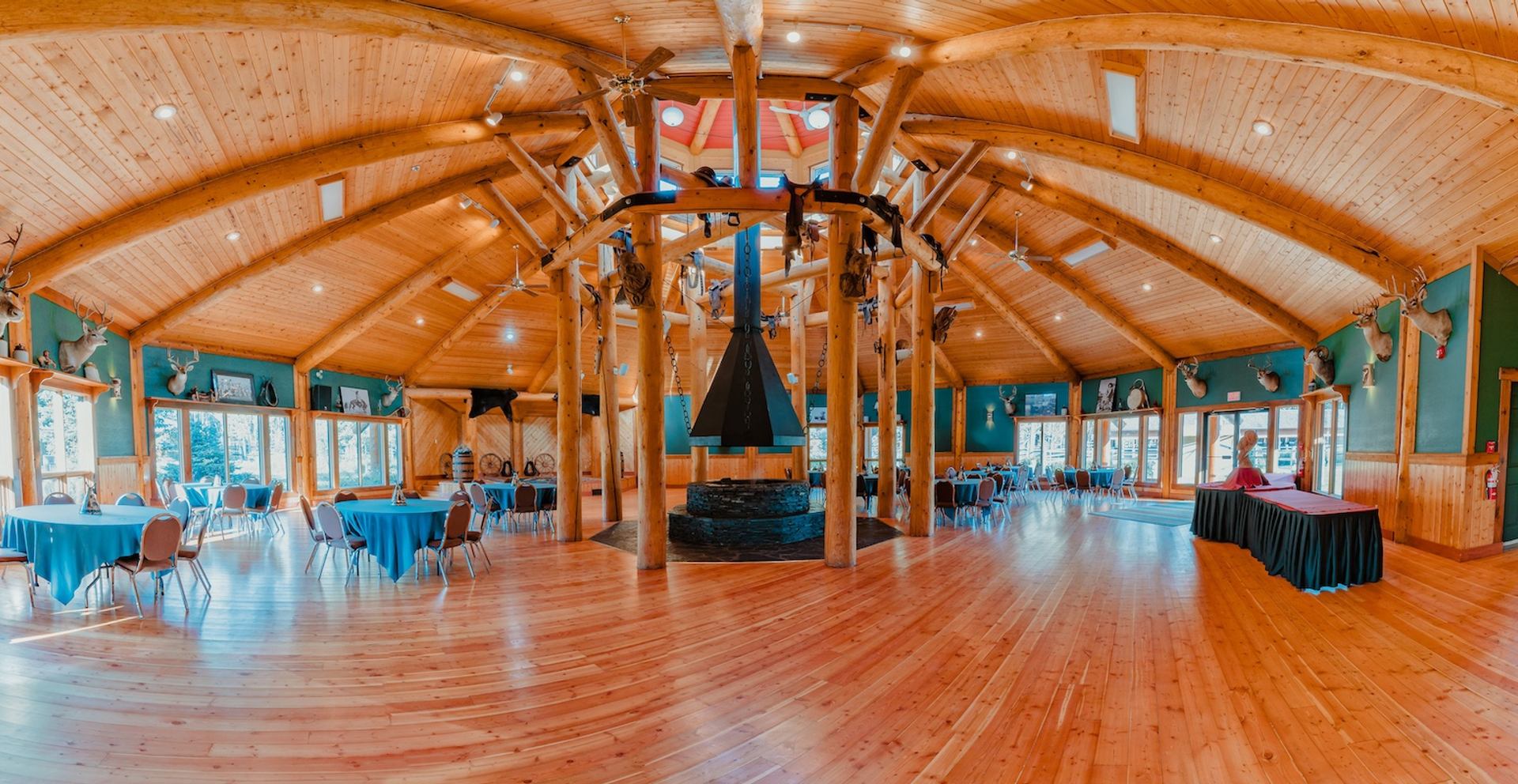 Spacious log‑beam event hall with round tables, tall ceilings, and central fireplace structure.