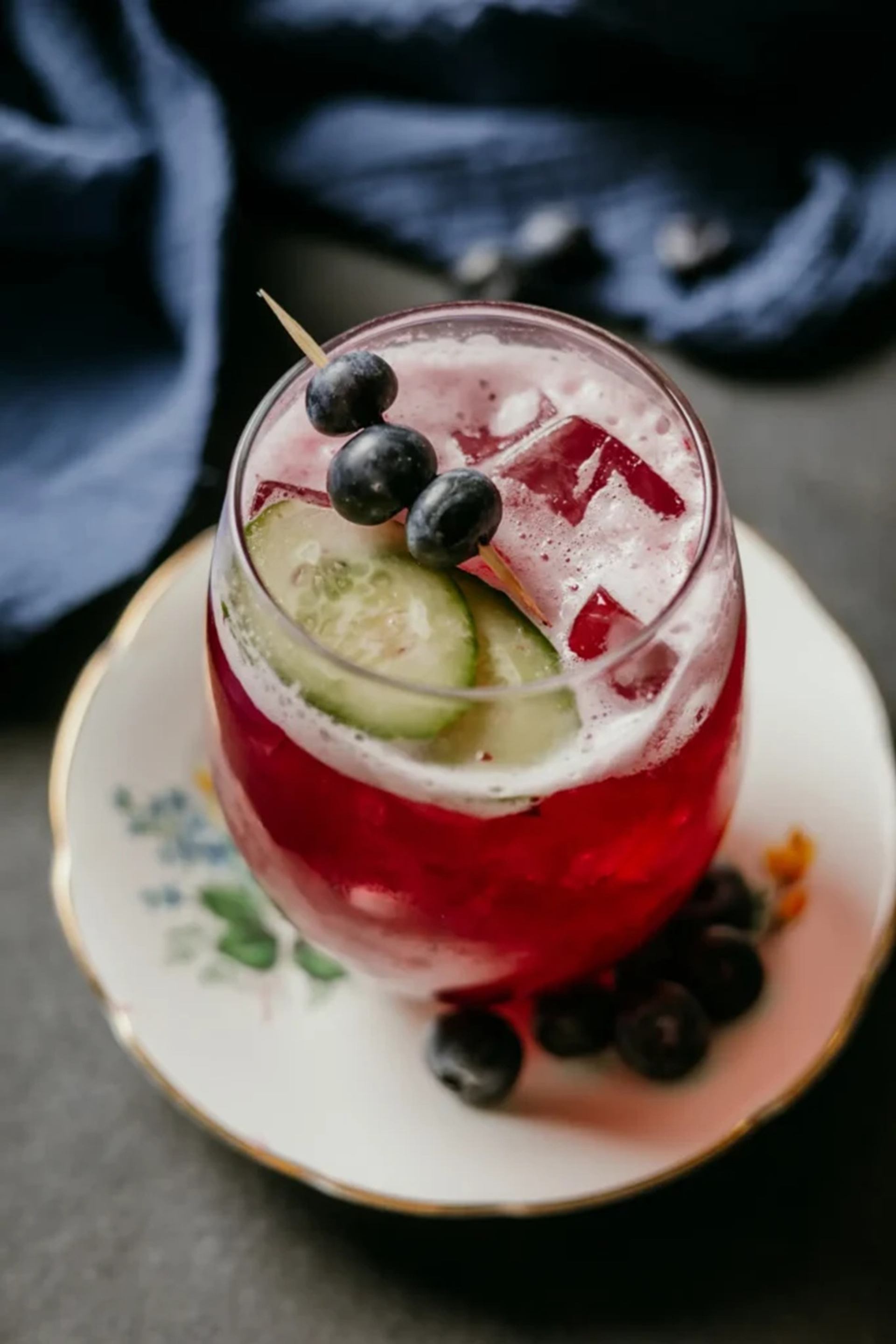 Red iced drink with cucumber slices and skewered blueberries on a floral saucer.