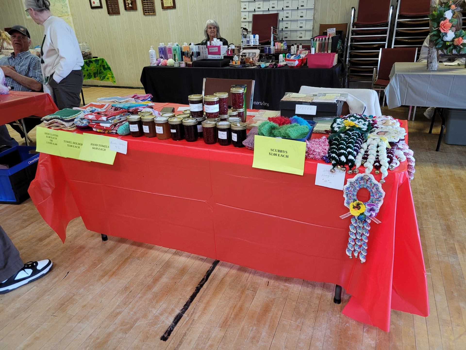 A red-covered table at a craft fair displays jars of preserves, fabric goods, and knitted items, with people browsing in the background.