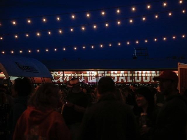 Crowd gathers under string lights in front of a saloon sign at night.
