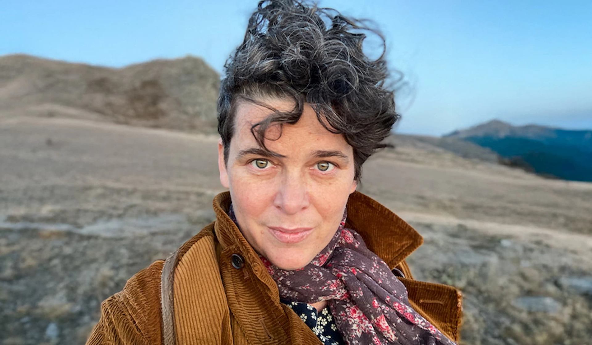 Person with short curly hair outdoors, wearing brown jacket and floral scarf.