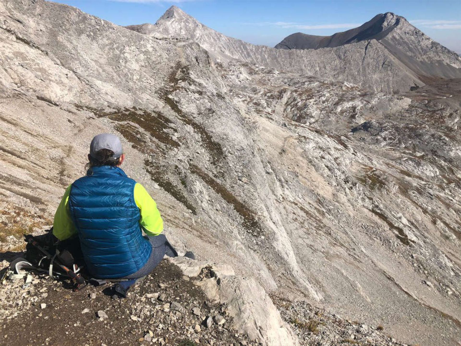 Hiker in blue vest sits on rocky slope overlooking rugged grey mountain peaks.