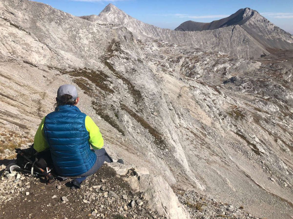 Hiker in blue vest sits on rocky slope overlooking rugged grey mountain peaks.