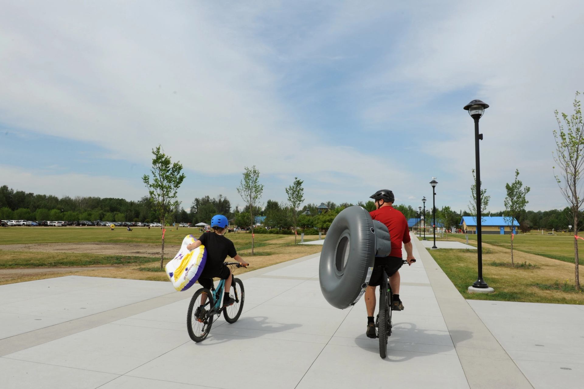 Two cyclists carry inflatable tubes and towels on a paved park path.