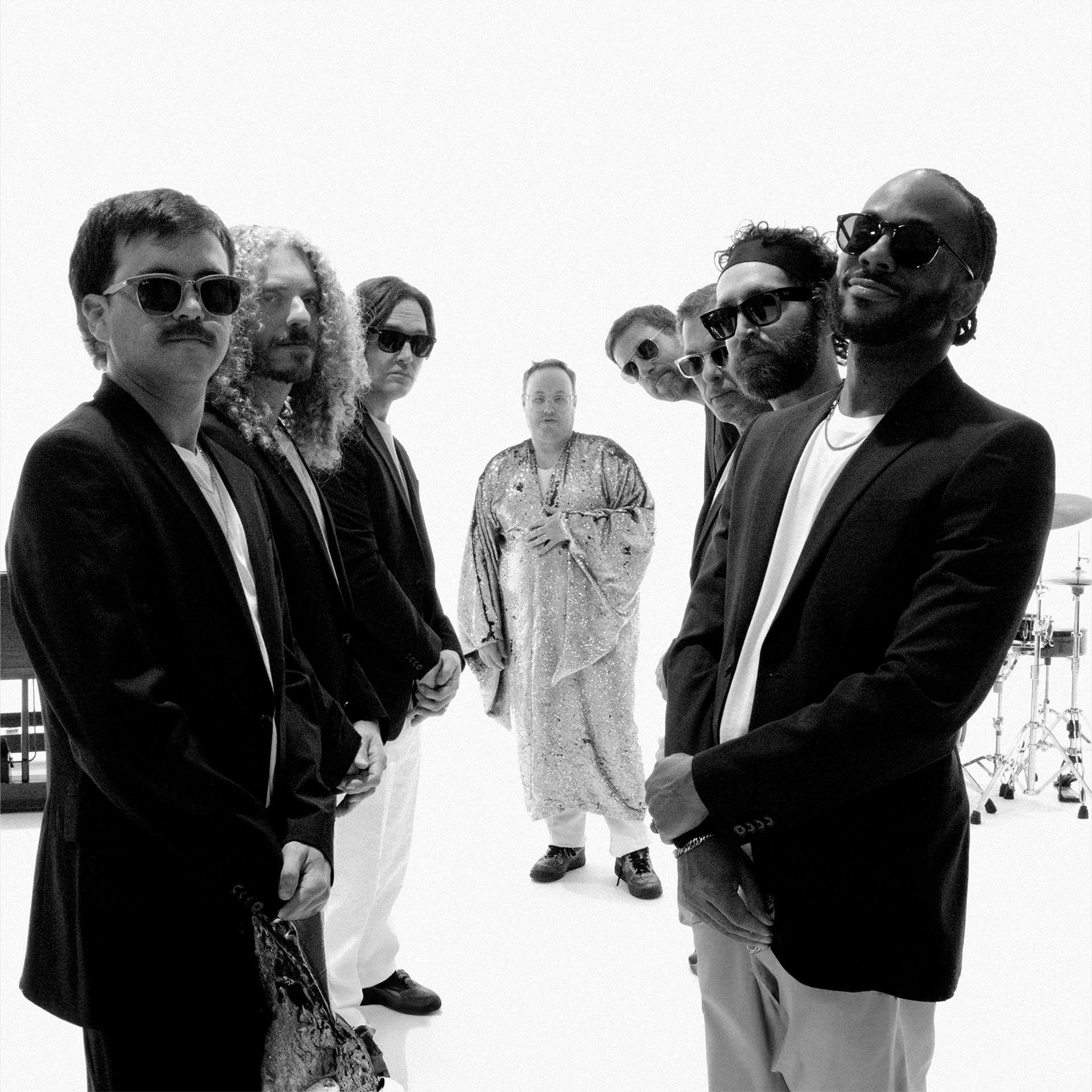 Musicians in suits and sunglasses stand around a robed figure in bright studio.