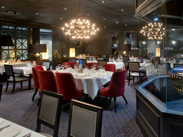 Restaurant dining room with round tables, white tablecloths, red chairs and chandeliers.