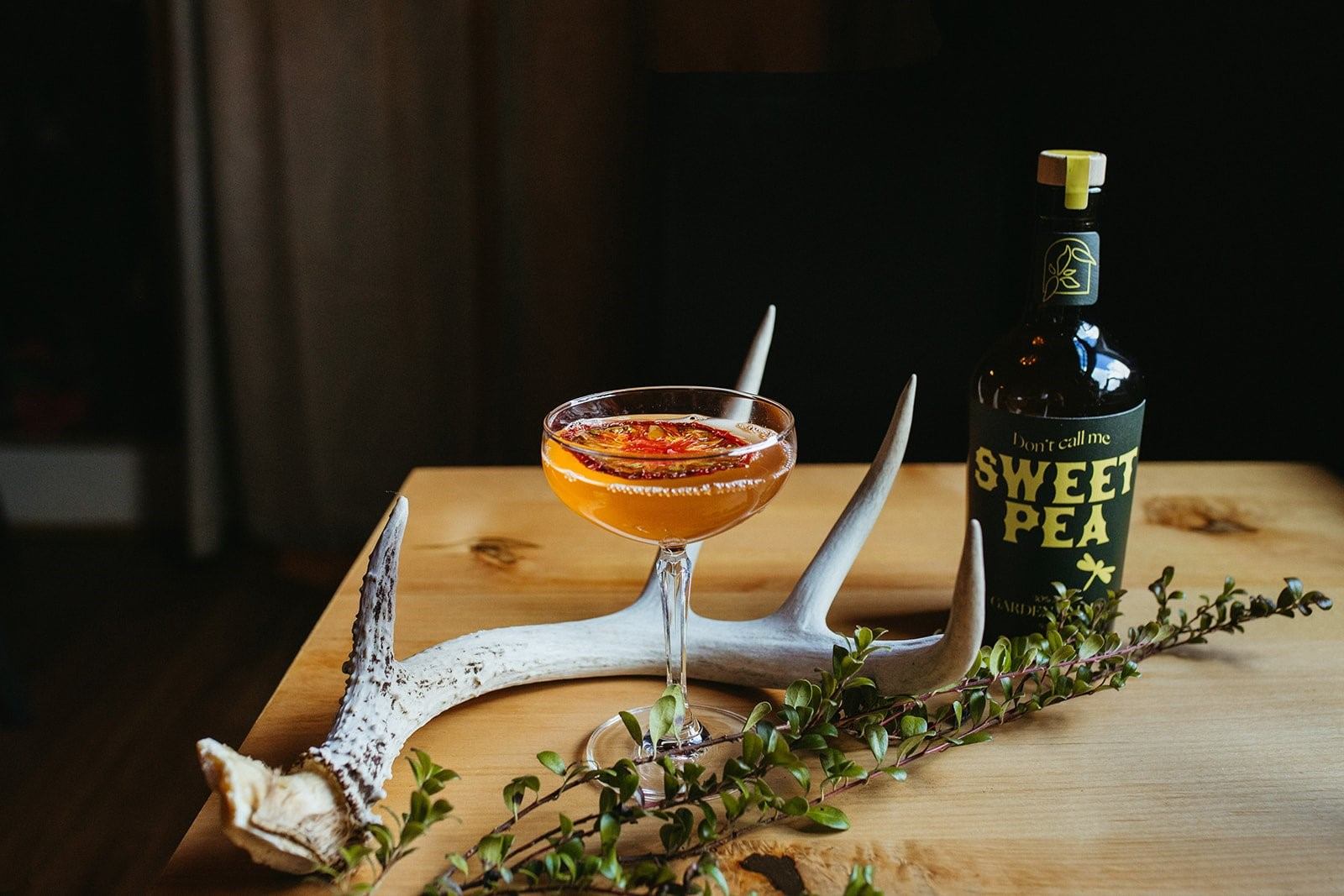 A cocktail in a glass with a Sweet Pea bottle, antlers, and greenery on a wooden table.
