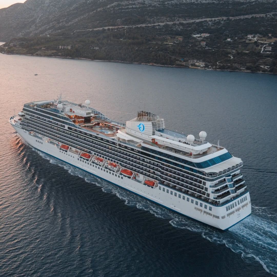 Large white cruise ship sails through calm water beside a mountainous coastline.