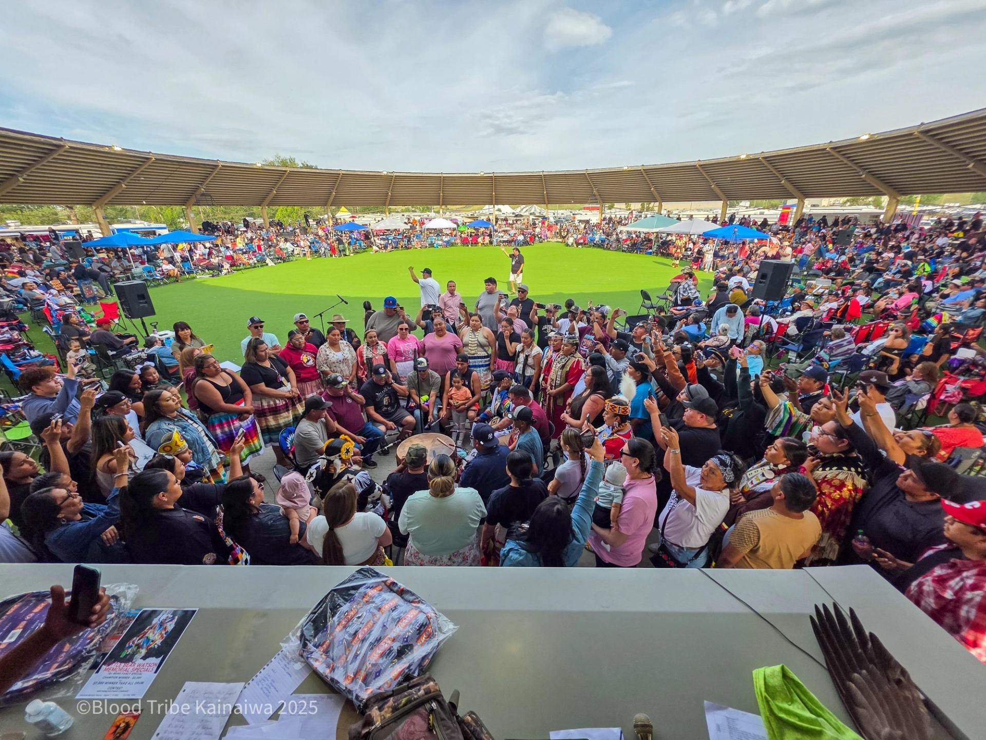 Large crowd gathers in covered arena, surrounding central drum group on green field.