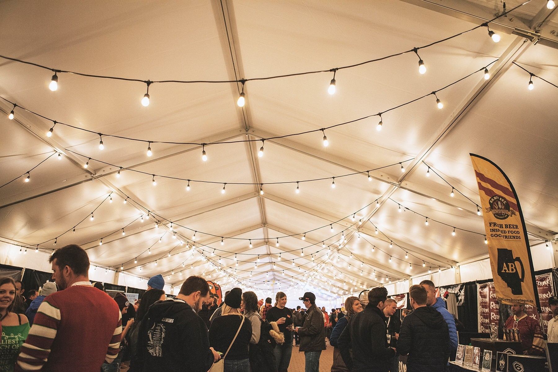 Indoor beer festival with string lights and lively crowd under a white tent.