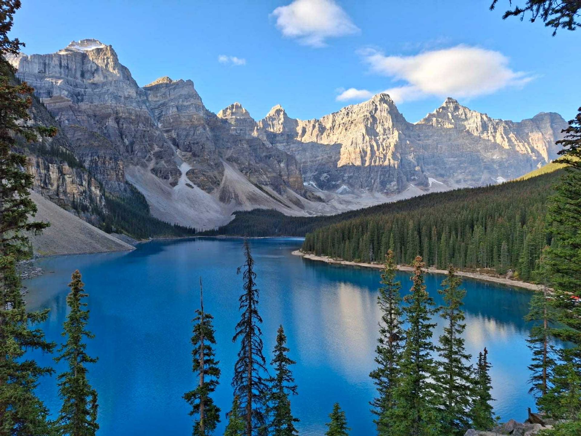 Blue mountain lake surrounded by tall peaks and evergreen forest under a bright sky.