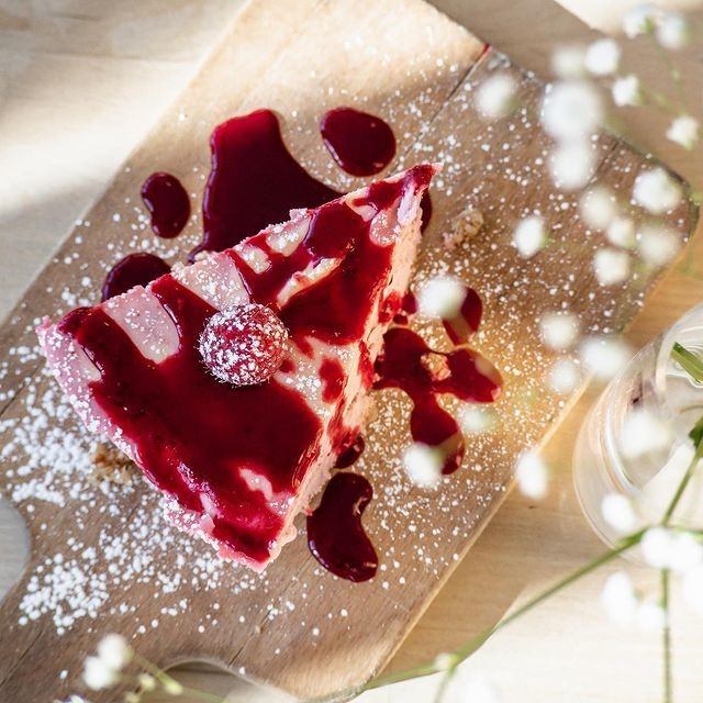 Slice of cake with berry sauce, powdered sugar, and floral garnish on wooden board.