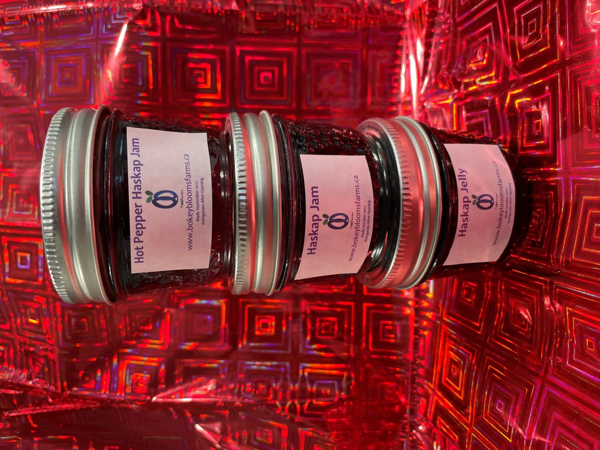 Three Bokey Blooms Farms haskap jam jars on reflective red geometric background.