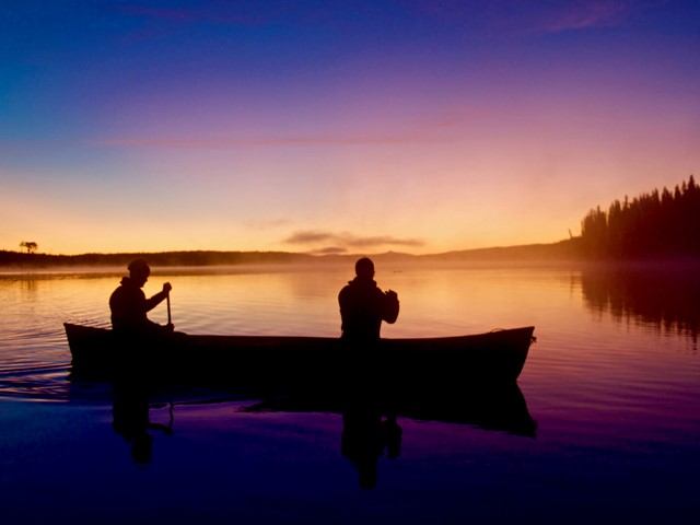 Two people paddling a canoe on a still lake at sunrise with vivid colors in the sky.