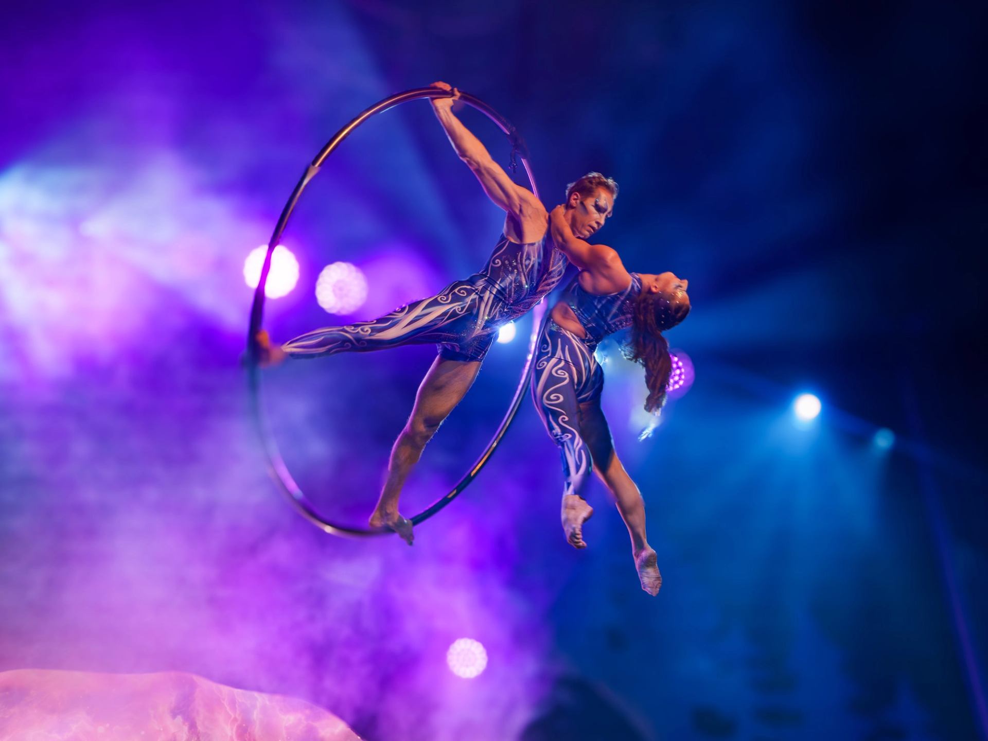 Aerial acrobats perform together inside a suspended hoop under purple and blue stage lights.