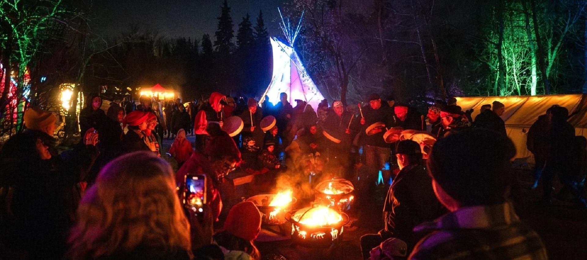 Group gathered around fire pits with colorful lights and a teepee at Flying Canoë Volant festival.