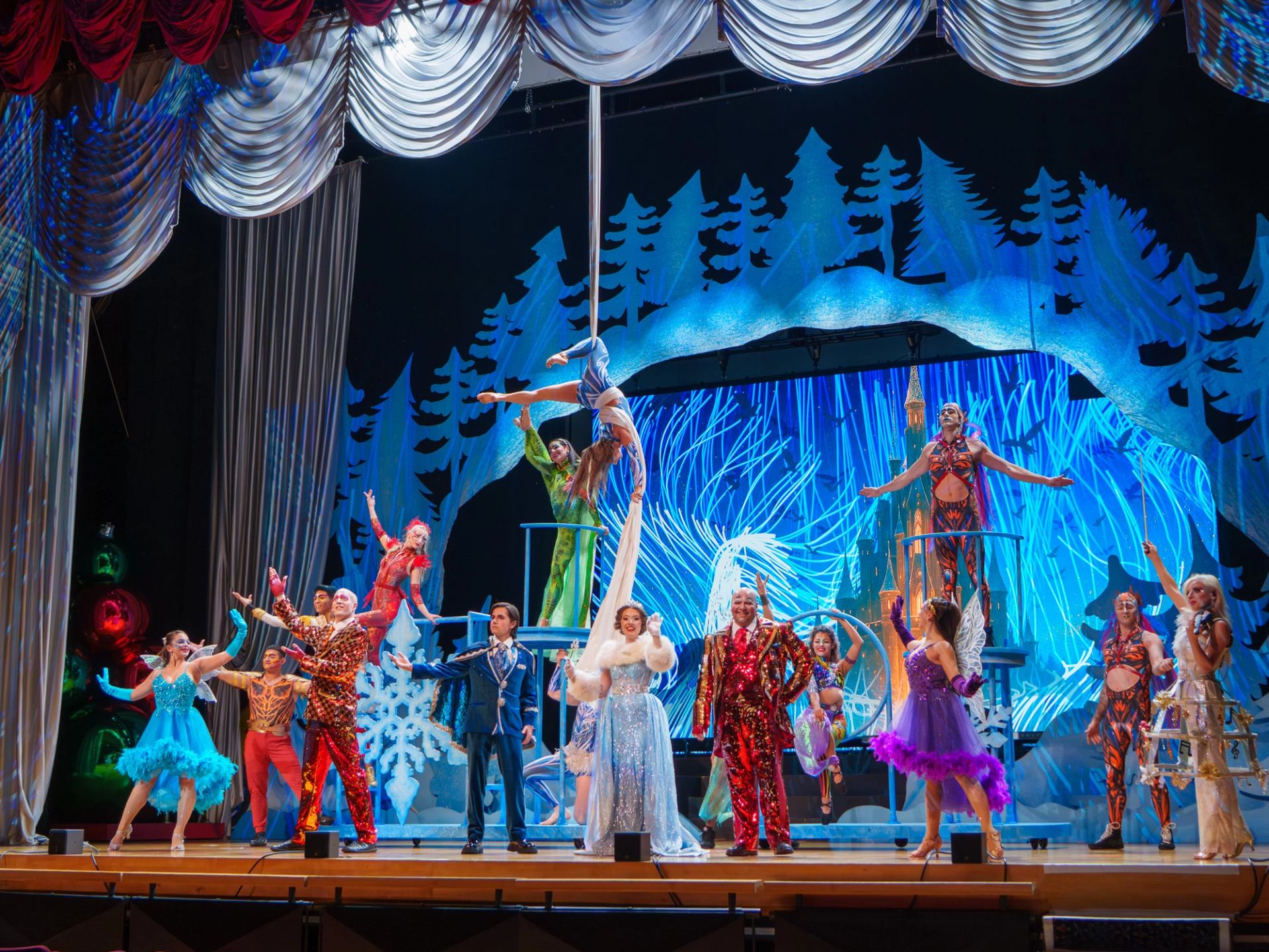 Costumed performers pose beneath an aerialist on a brightly lit theatre stage.