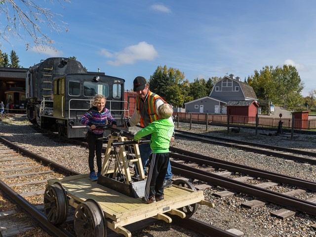 Three people on a handcar with train and buildings in the background.