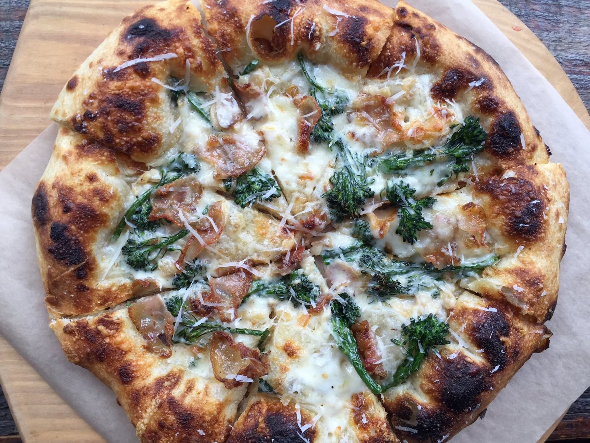 A wood-fired pizza is topped with melted cheese, broccolini, cured meat, and shaved parmesan.