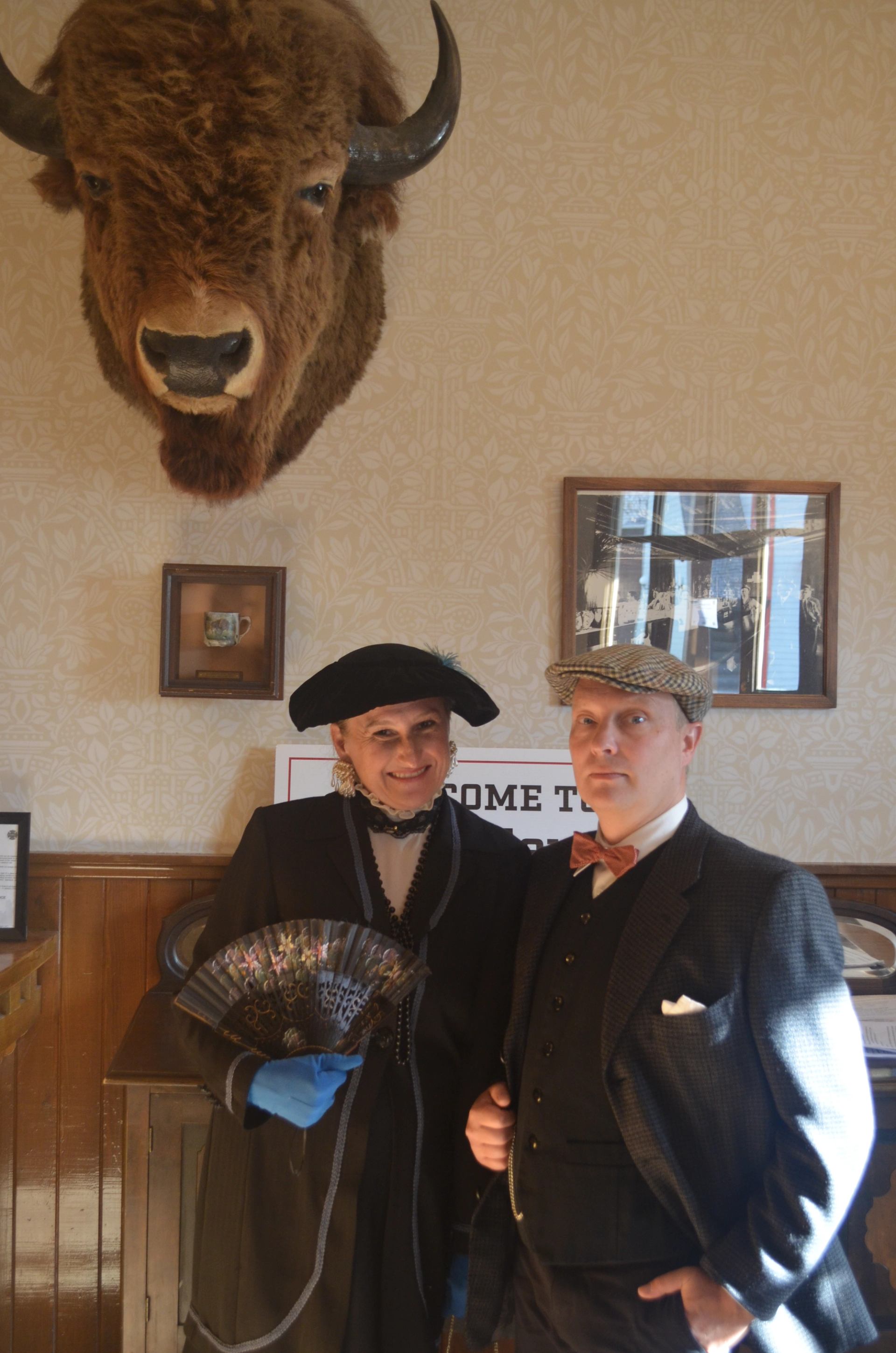 People in vintage clothing pose beneath a mounted bison head and framed pictures.