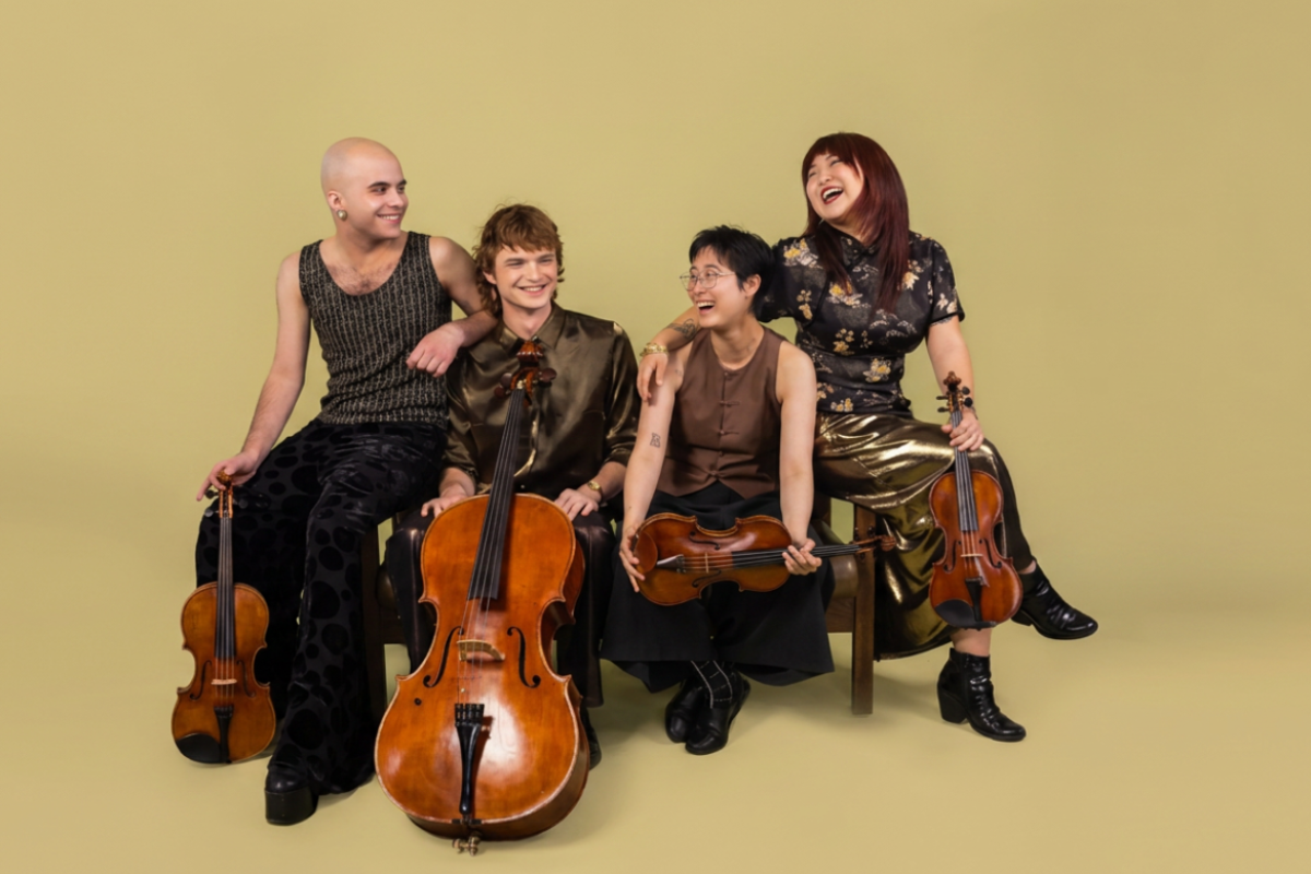 String quartet musicians sitting together with violins and cellos against a plain yellow background.