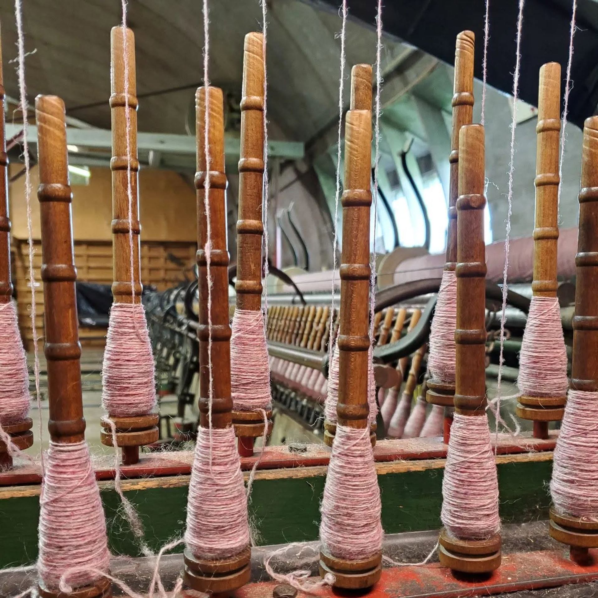 Wooden spindles with pink yarn on a textile machine inside an industrial workshop.