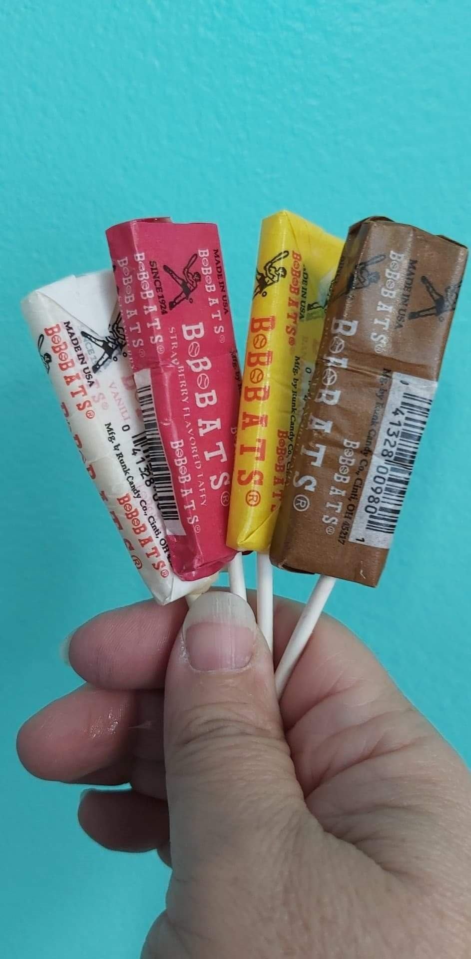 Hand holding four assorted Reed’s candy sticks against a teal background.