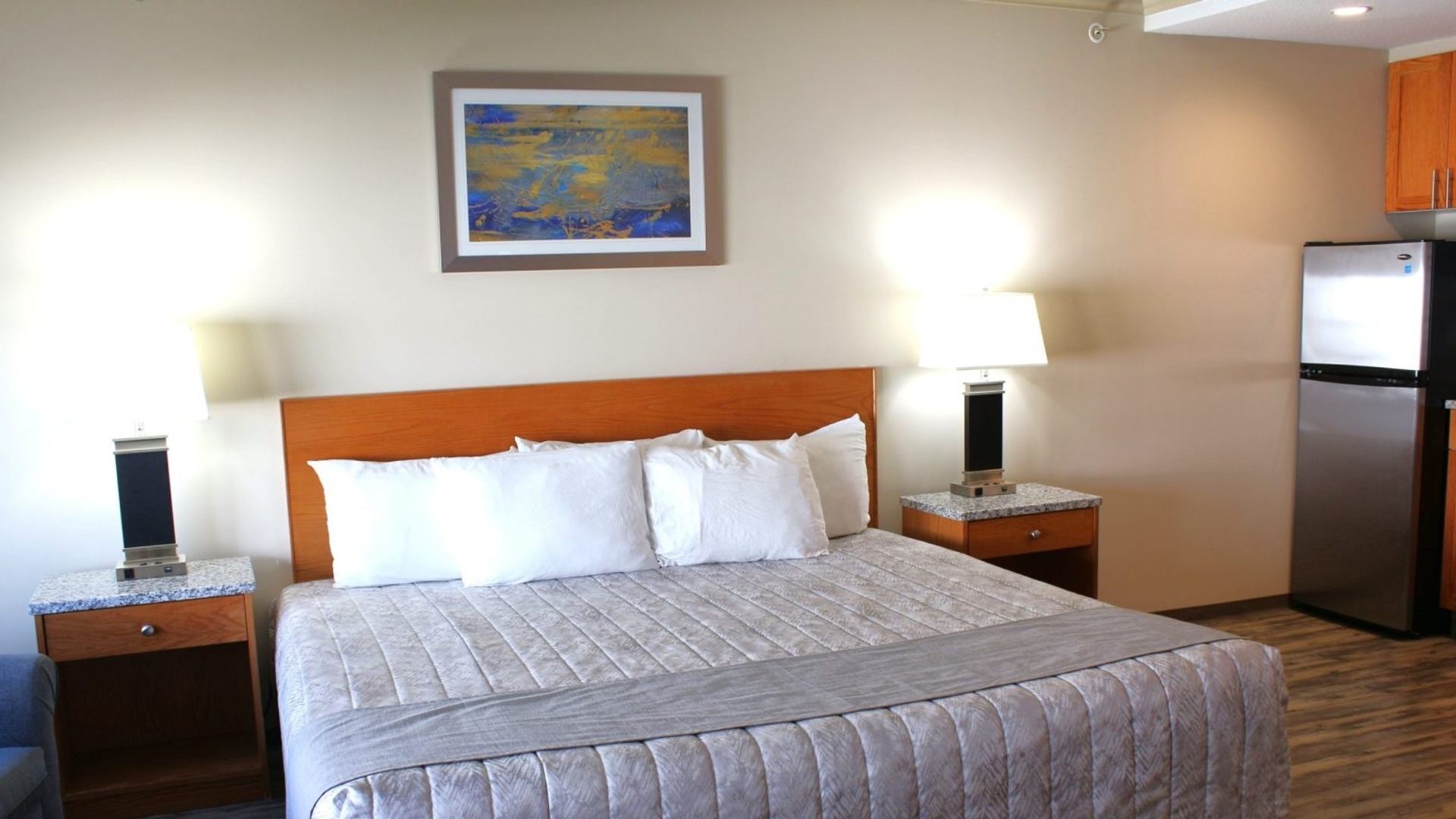 Modern motel room with king bed, lamps, artwork, and mini-fridge.
