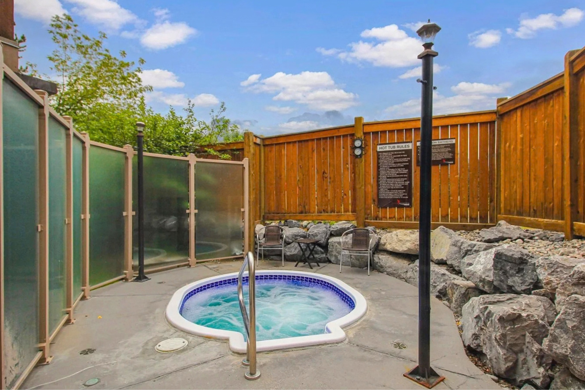 Outdoor hot tub with privacy screens, wooden fencing, rocks and patio chairs.
