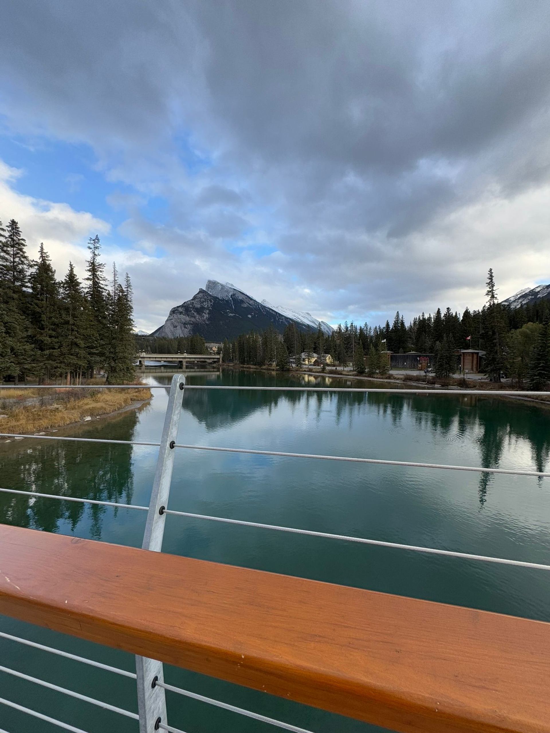 Mountain peak and evergreen trees reflected in calm river beside bridge railing.