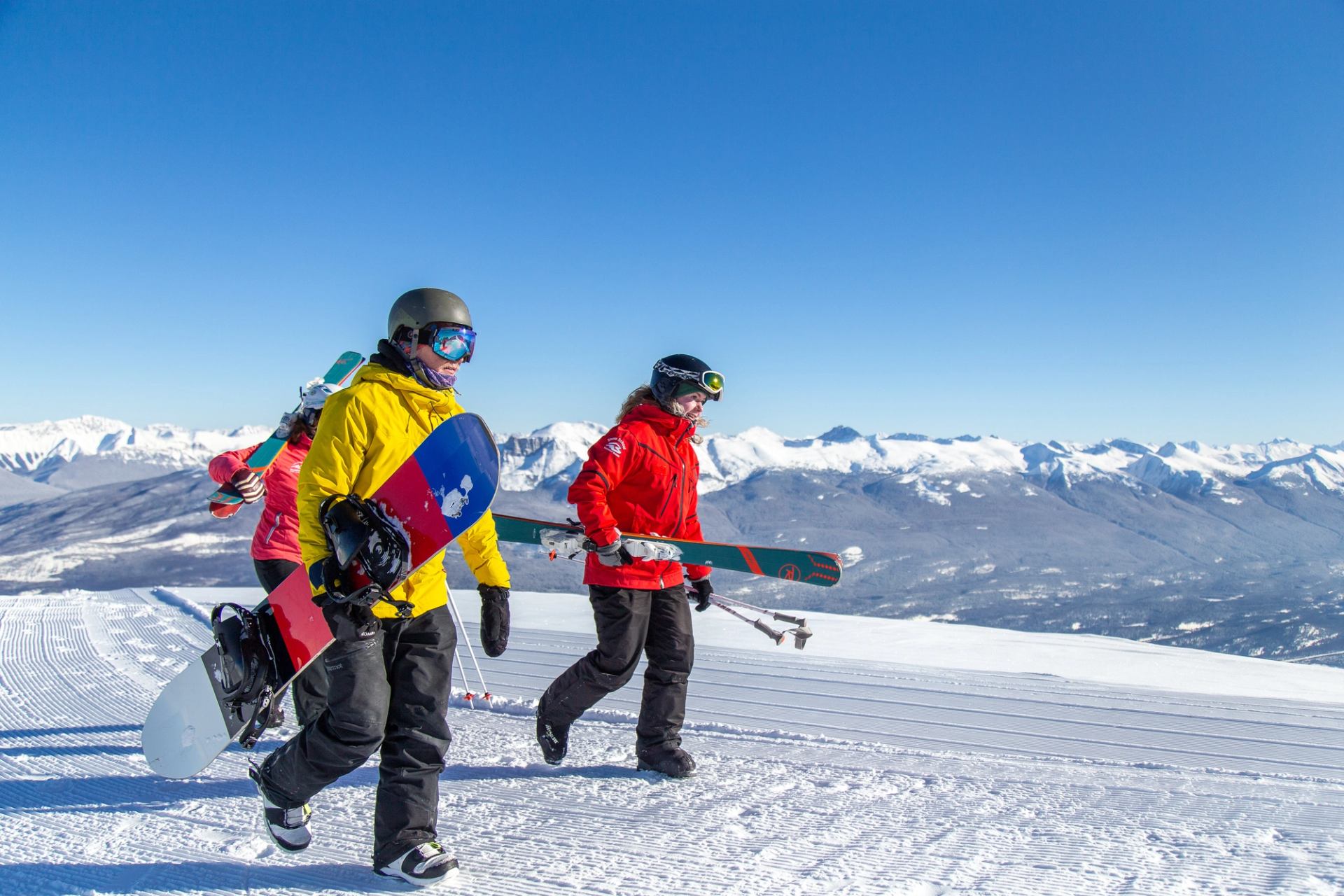 Skiers and snowboarders walking across a snowy ridge with clear skies and mountain views.